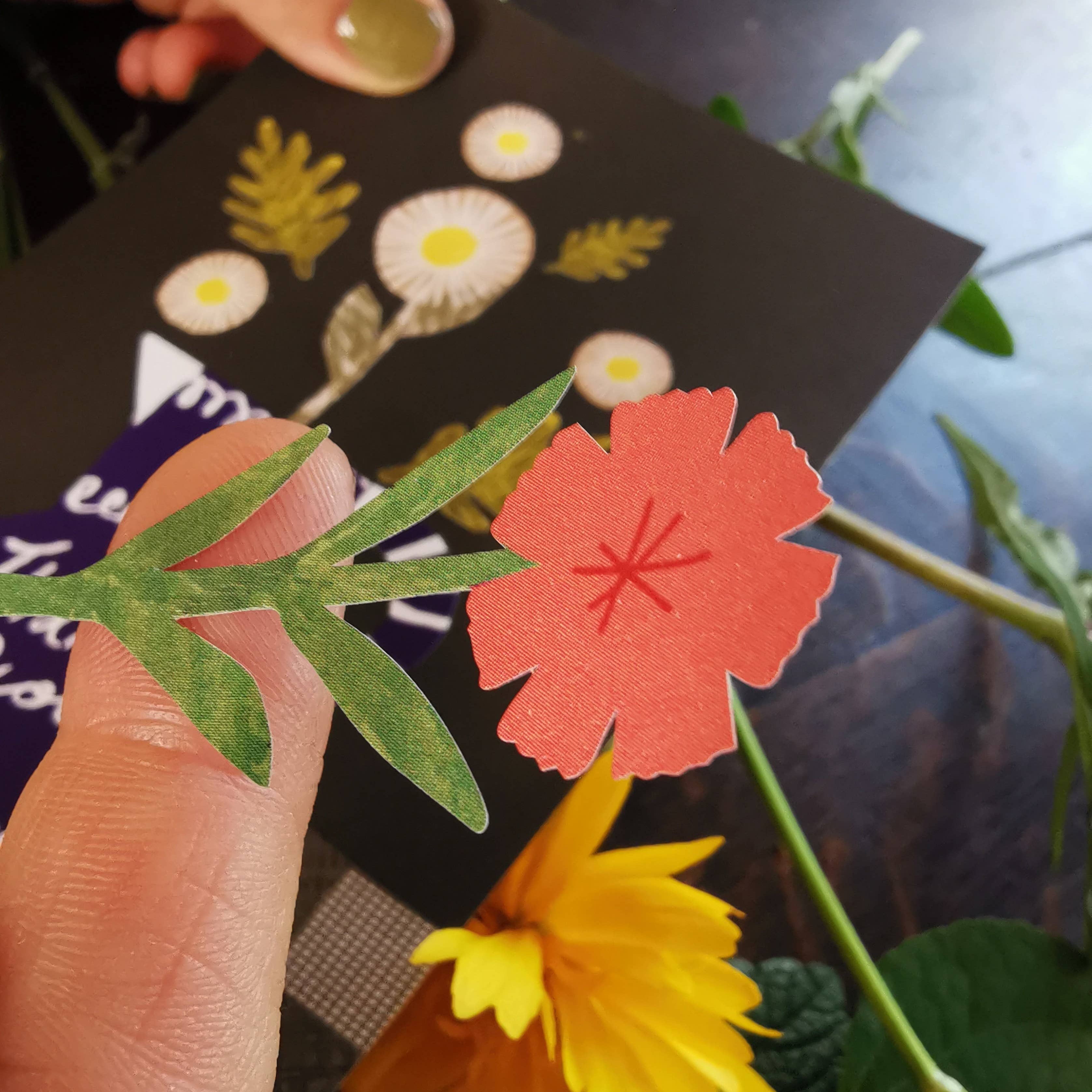Hand holding a black card with floral designs and a red flower cutout, surrounded by green leaves and a yellow flower.
