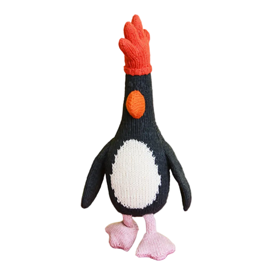 A crocheted penguin doll with a red comb, black and white body, and pink feet, displayed on a yellow and orange background.