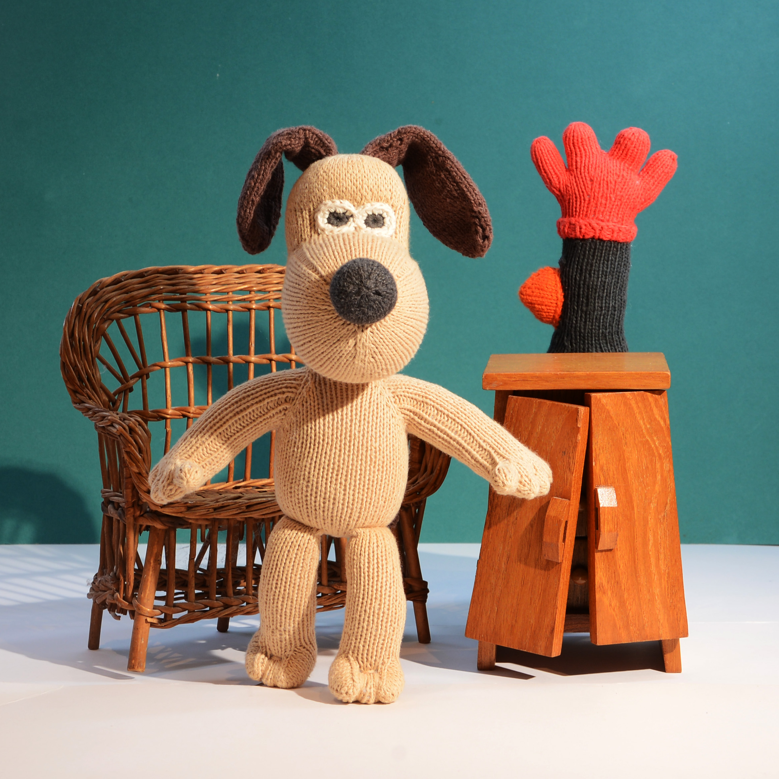 A knitted dog plush toy with floppy dark brown ears stands in front of a teal studio backdrop, next to a small wooden cabinet and a red-and-black glove prop.