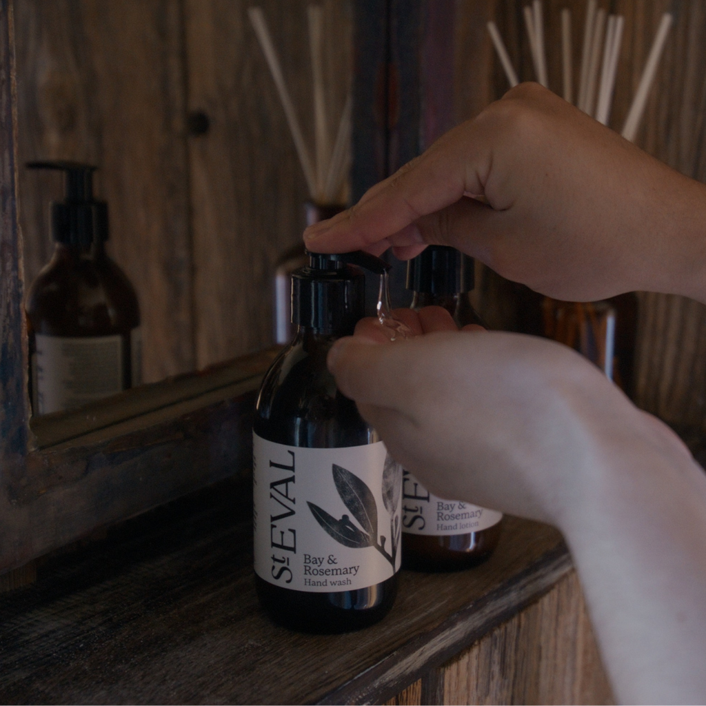 Right hand pumping the hand wash liquid into left hand. Hand wash bottle sits on a wooden surface with a mirror behind, reflecting the soap bottle.