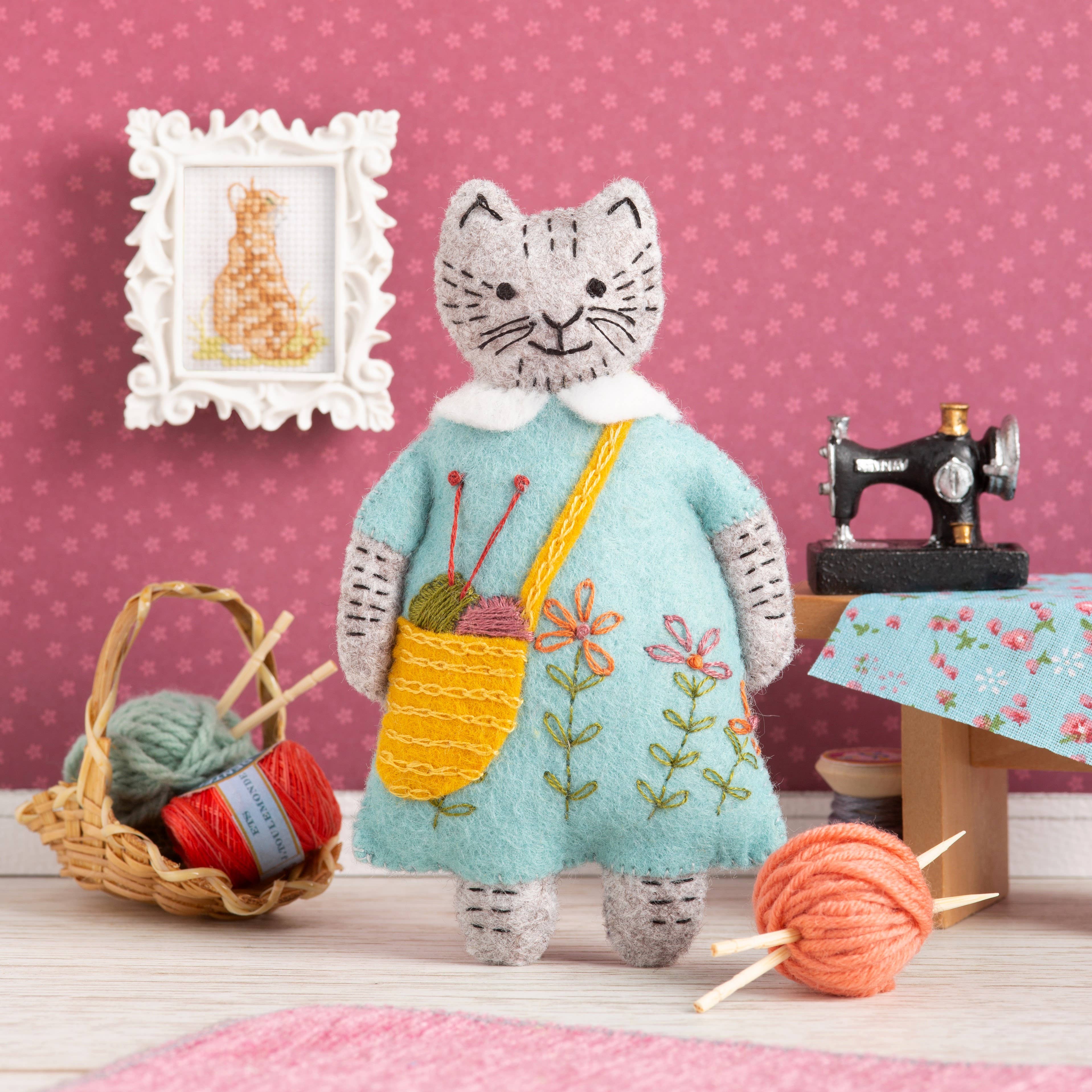 Cat-shaped plush toy in a room with sewing equipment and yarn on a pink polka dot wall background.