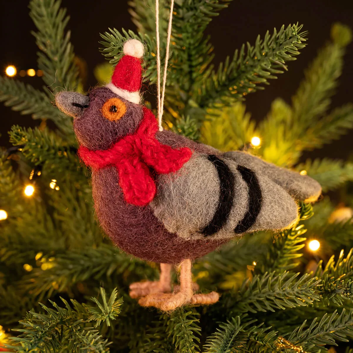 A felted wool Christmas ornament shaped like a pigeon, wearing a red and white Santa hat and a red scarf, hanging on a green Christmas tree with warm string lights.