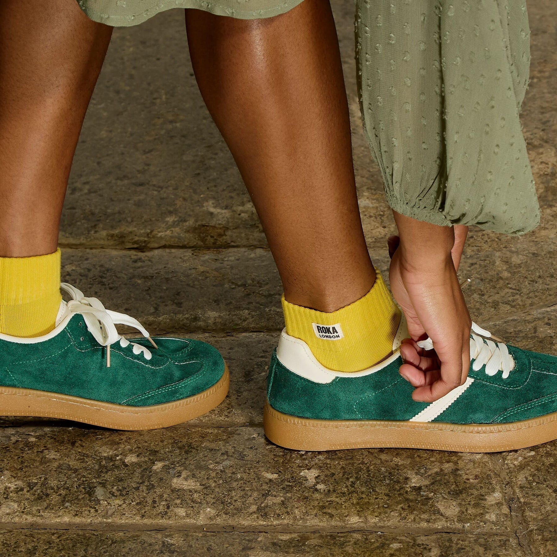 Person wearing green sneakers with yellow socks and a visible brand logo, standing on a stone surface.