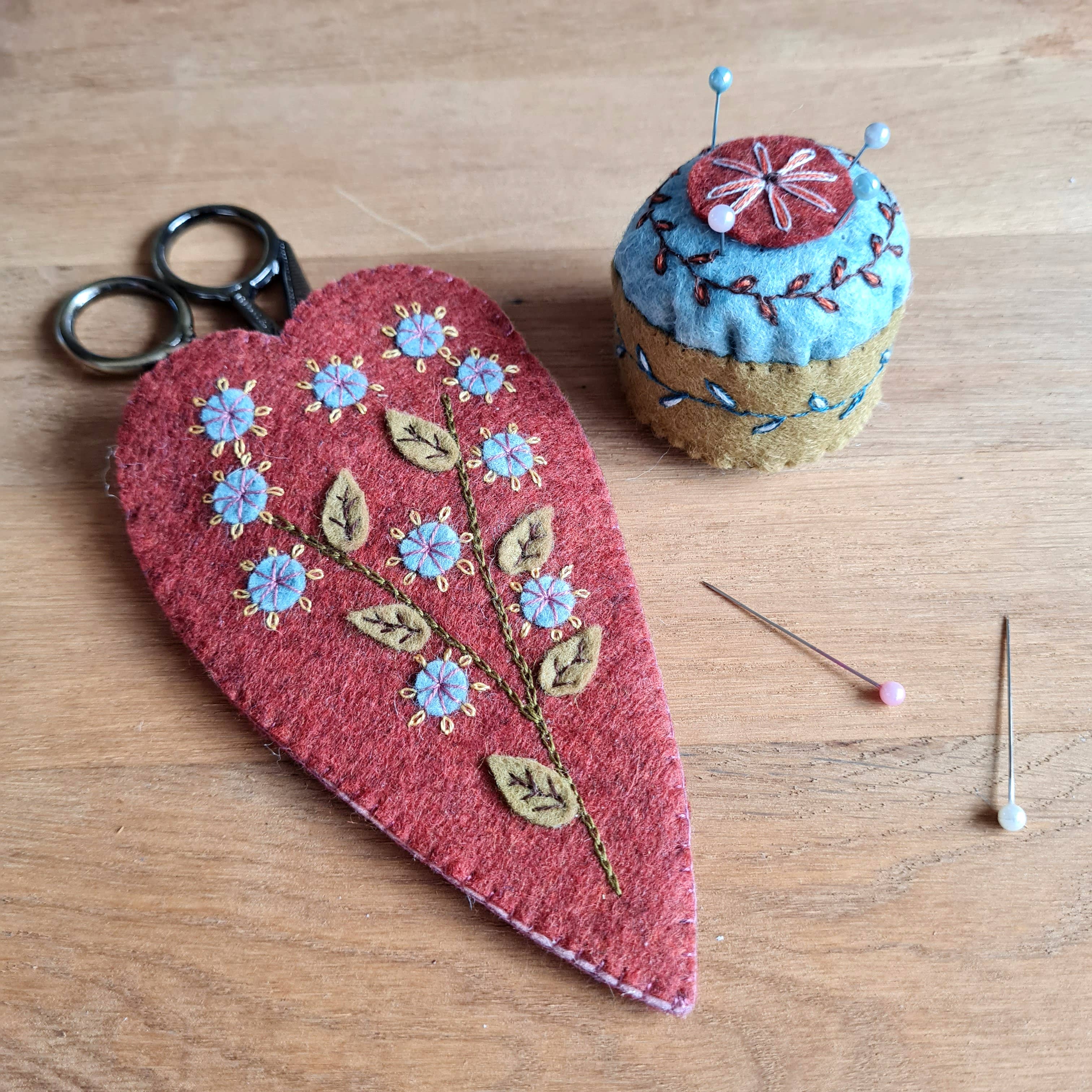 Embroidered red heart-shaped scissors pouch, colourful mini pincushion with botanical design, and sewing tools on a wooden surface.