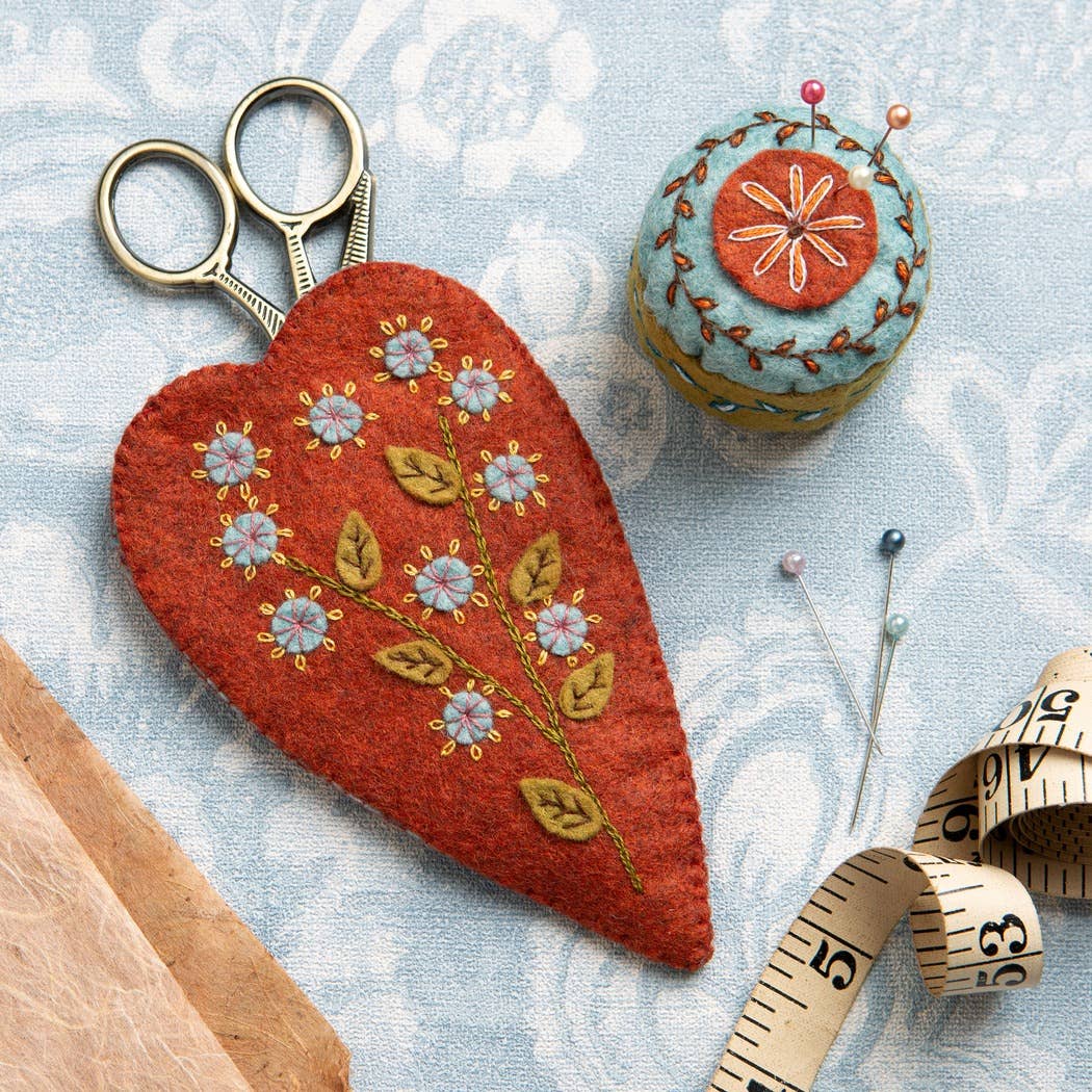Embroidered red heart-shaped scissors pouch, colourful mini pincushion with botanical design, and sewing tools on a patterned blue fabric surface.