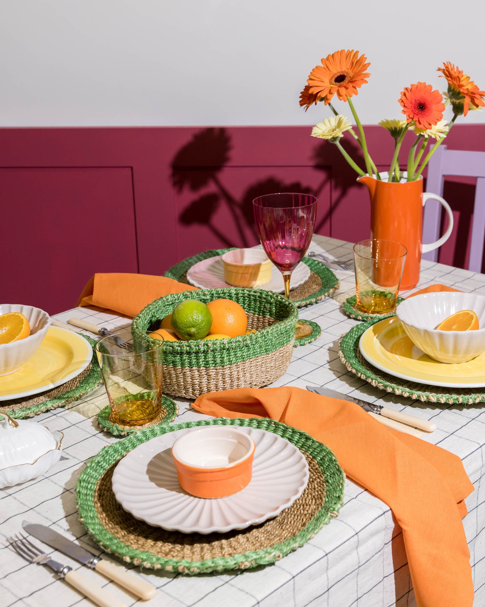 Dining table set with orange and green tableware, flowers, and fruit on a white tablecloth.