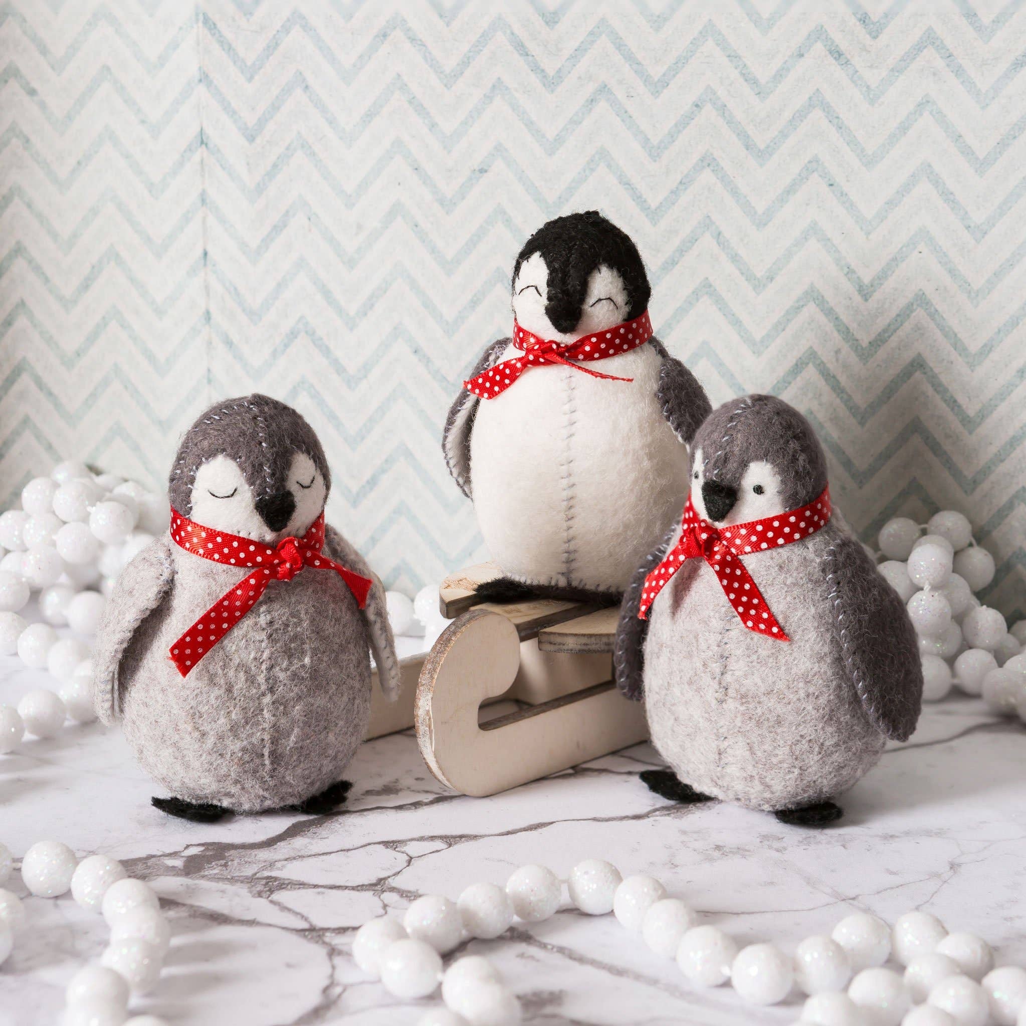 Three felt penguins with red polka dot bows on a marble surface with decorative white balls and a chevron-patterned wall.