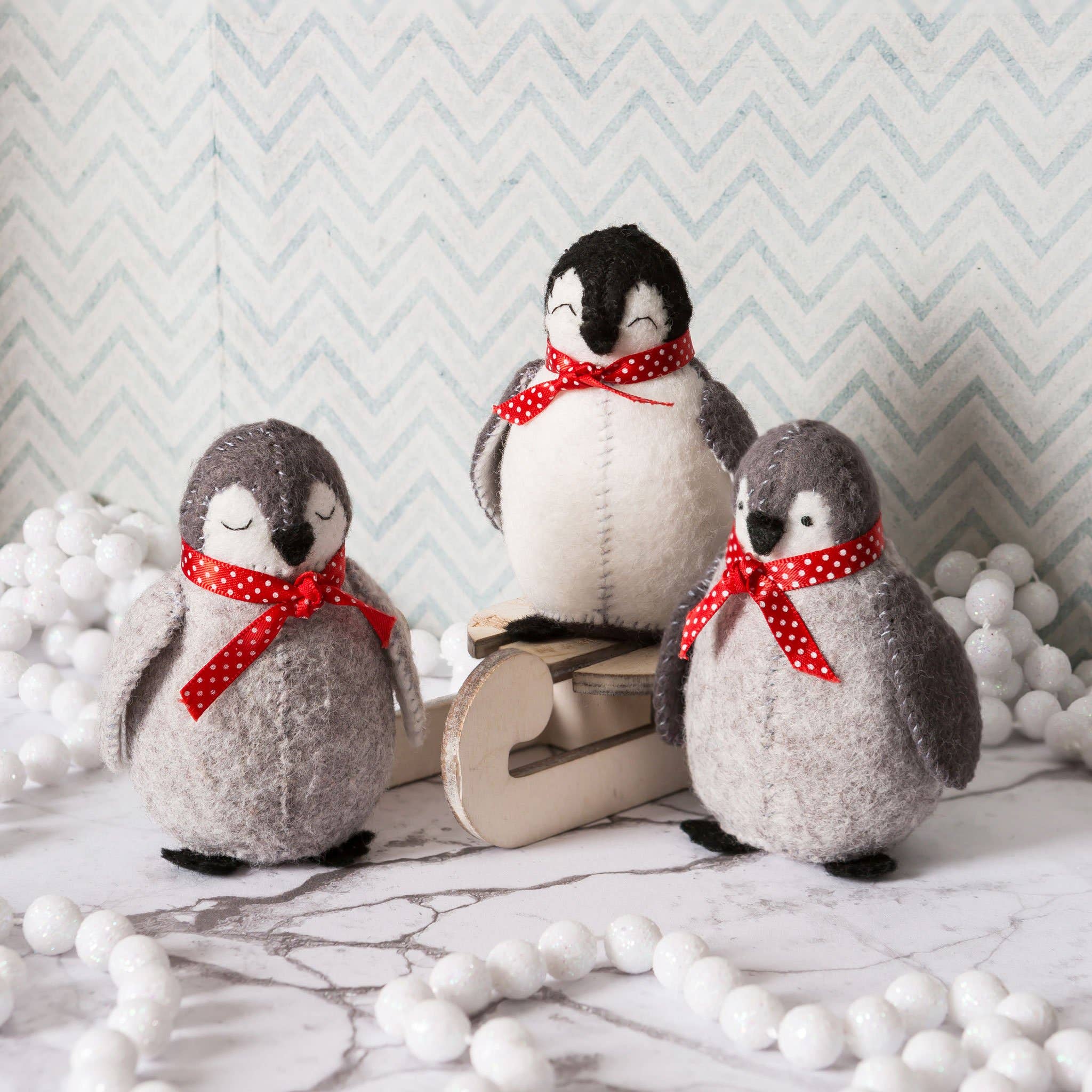Three felt penguins with red polka dot bows on a marble surface with decorative white balls and a chevron-patterned wall.