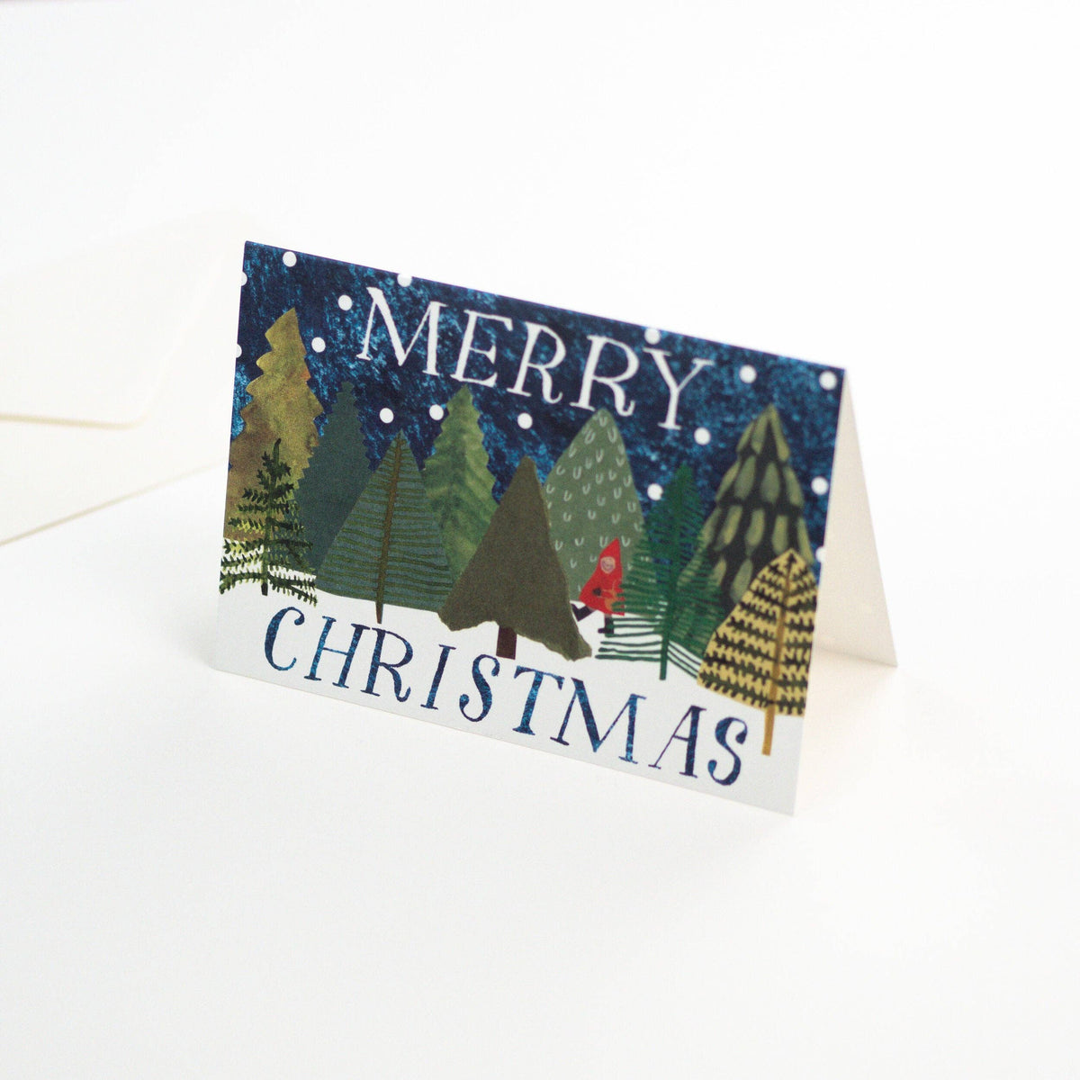 A folded Christmas greeting card standing upright on a white surface. The card features a winter forest scene with pine trees, snow, and the words “MERRY CHRISTMAS” in blue lettering.