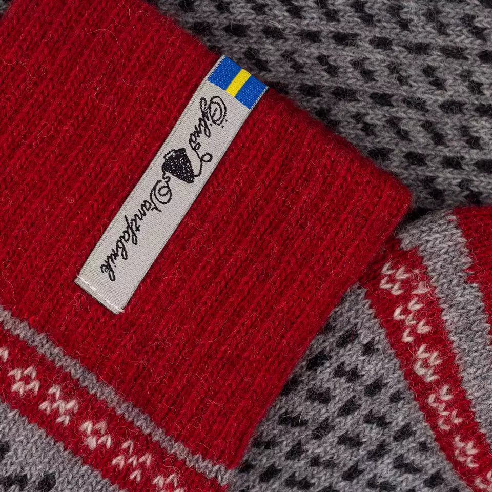 Close-up of a red knit sock with a white brand tag and a small Swedish flag, laid over a gray and black patterned knit textile.