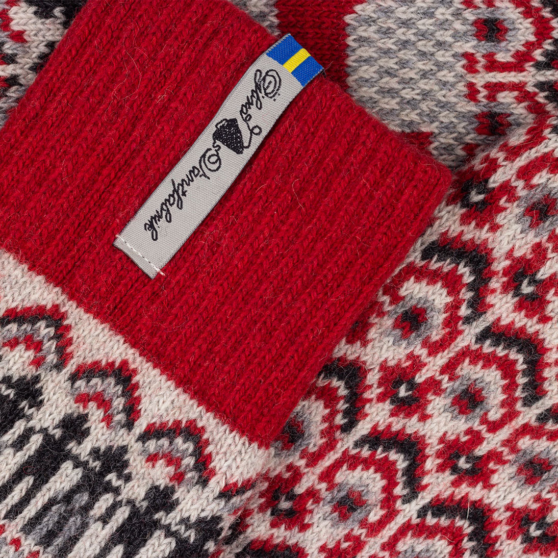Close-up of a knitted socks showing a red ribbed cuff with a white tag alongside a red, gray, and black geometric pattern.