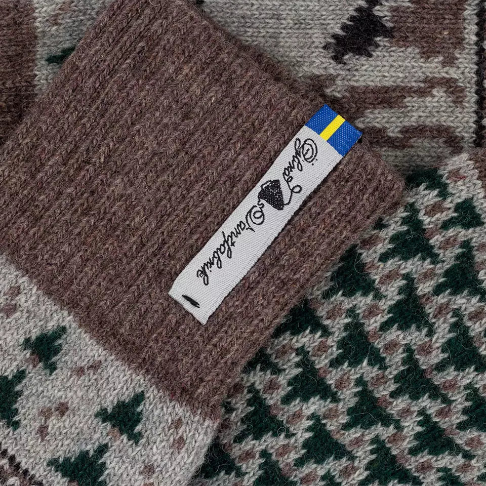 Close-up of a knitted sock with a brown ribbed cuff featuring a white tag that reads 'Guld Handgross' with a small blue and yellow accent, over a patterned scarf in brown, beige, and green.