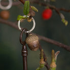 A close-up of a metal keyring with a carved acorn pendant hanging from a dark metal key, set against a blurred natural background with leaves and a red berry.