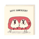 A square greeting card with a cream background featuring the text 'HAPPY ANNIVERSARY' above an illustration of two sheep sitting on a pink couch, with a matching cream envelope behind it.