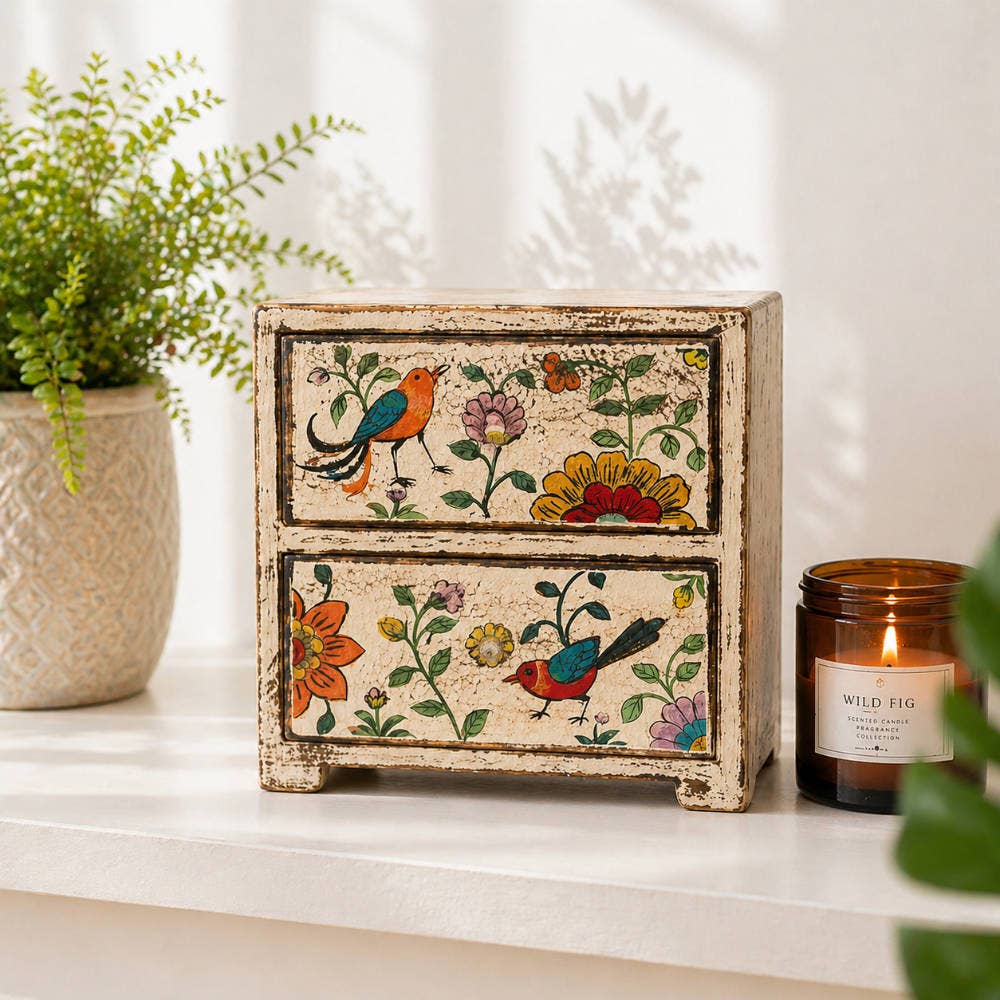 Decorative wooden box with floral and bird patterns on a white surface with a candle and plant in the background.