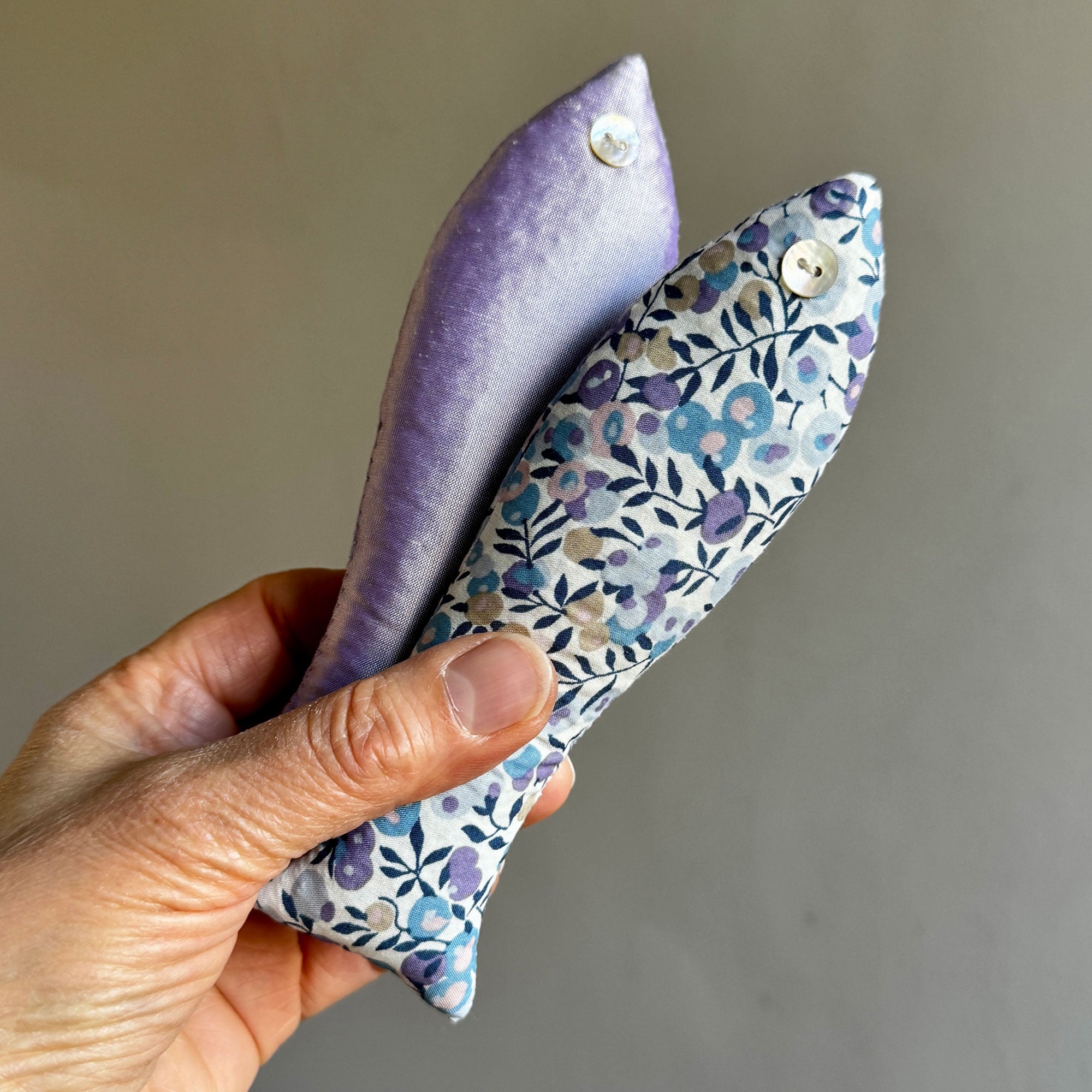 Hand holding two cotton lavender sachets with floral patterns on a plain background