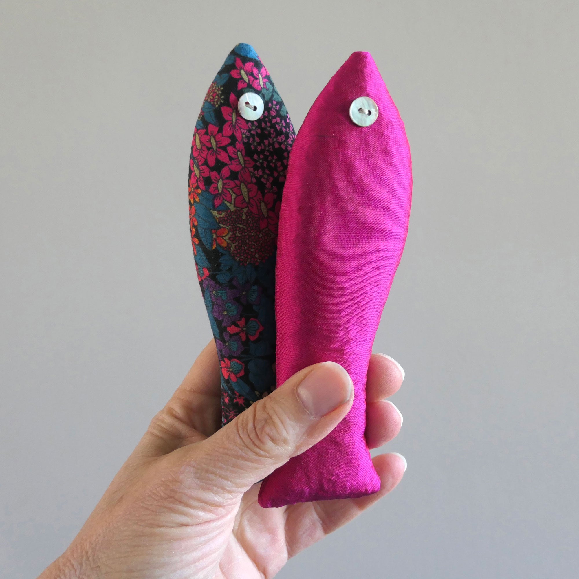 Hand holding two fabric fish sachets, one pink and one with floral pattern, against a plain background.