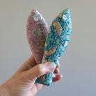 Hand holding two floral-patterned fish-shaped  sachets in Strawberry Thief Liberty Fabric against a plain background