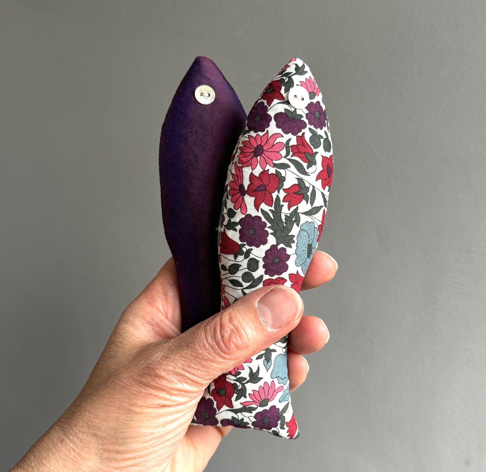 Hand holding two cotton lavender sachets, one purple and one with floral pattern, against a grey background.