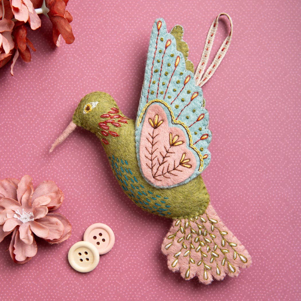 Embroidered felt hummingbird decoration in greens, blues and pinks shown on a pink patterned background with flowers and buttons around the bird. 