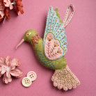 Embroidered felt hummingbird decoration in greens, blues and pinks shown on a pink patterned background with flowers and buttons around the bird. 