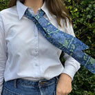 Person wearing a white shirt with a blue floral tie over one shoulder, standing against a green leafy background.