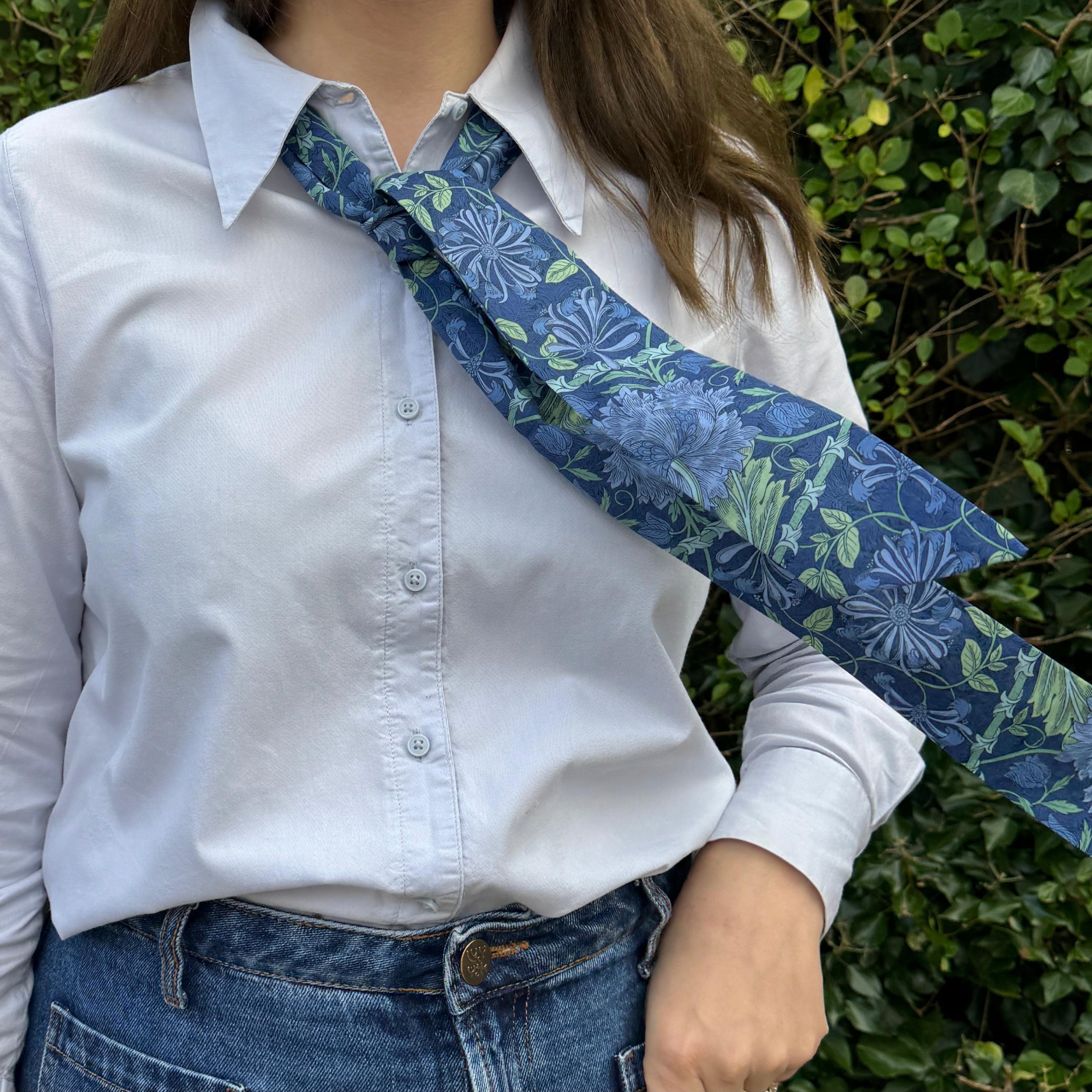 Person wearing a white shirt with a blue floral tie over one shoulder, standing against a green leafy background.