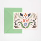 A greeting card with a  botanical illustrations, placed in front of a green envelope on a light background. The card features the word “JOY” in orange-red lettering with decorative leaves and small floral motifs around it.