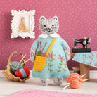 Cat-shaped plush toy in a room with sewing equipment and yarn on a pink polka dot wall background.