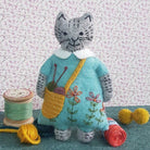 Felt grey cat toy wearing a blue embroidered dress and knitting bag, on a floral background with craft and knitting props around them. 