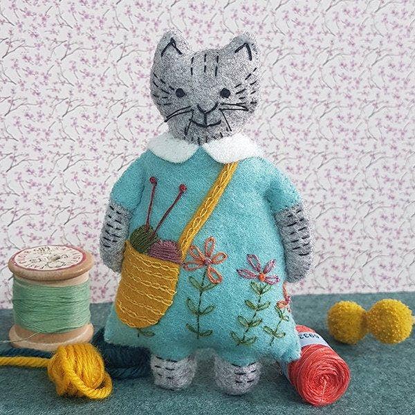 Felt grey cat toy wearing a blue embroidered dress and knitting bag, on a floral background with craft and knitting props around them. 