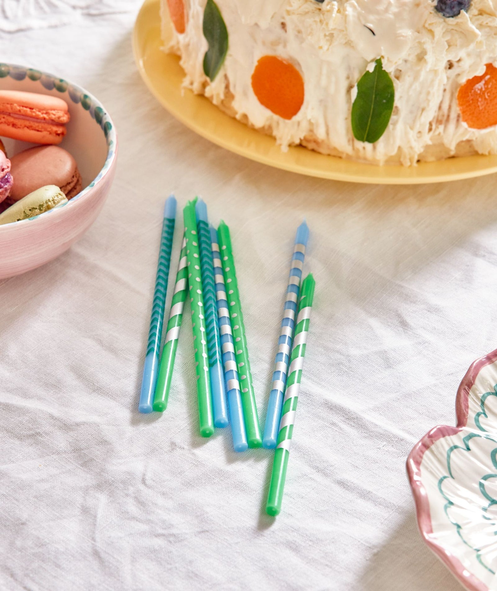 Colorful birthday candles on a white surface with a cake and macarons in the background.