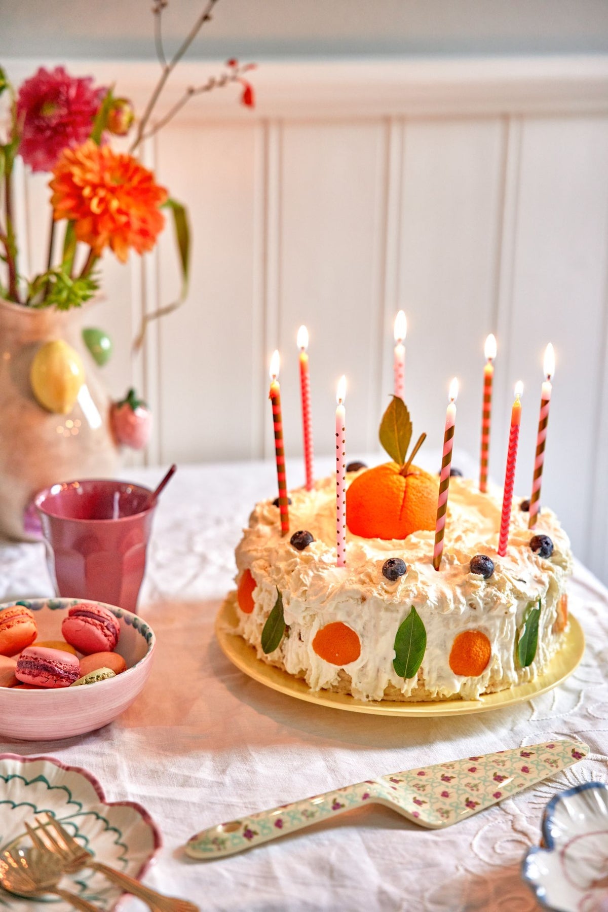 Birthday cake with lit candles on a table with flowers and a cup in the background