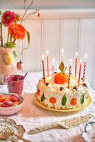 Birthday cake with lit candles on a table with flowers and a cup in the background