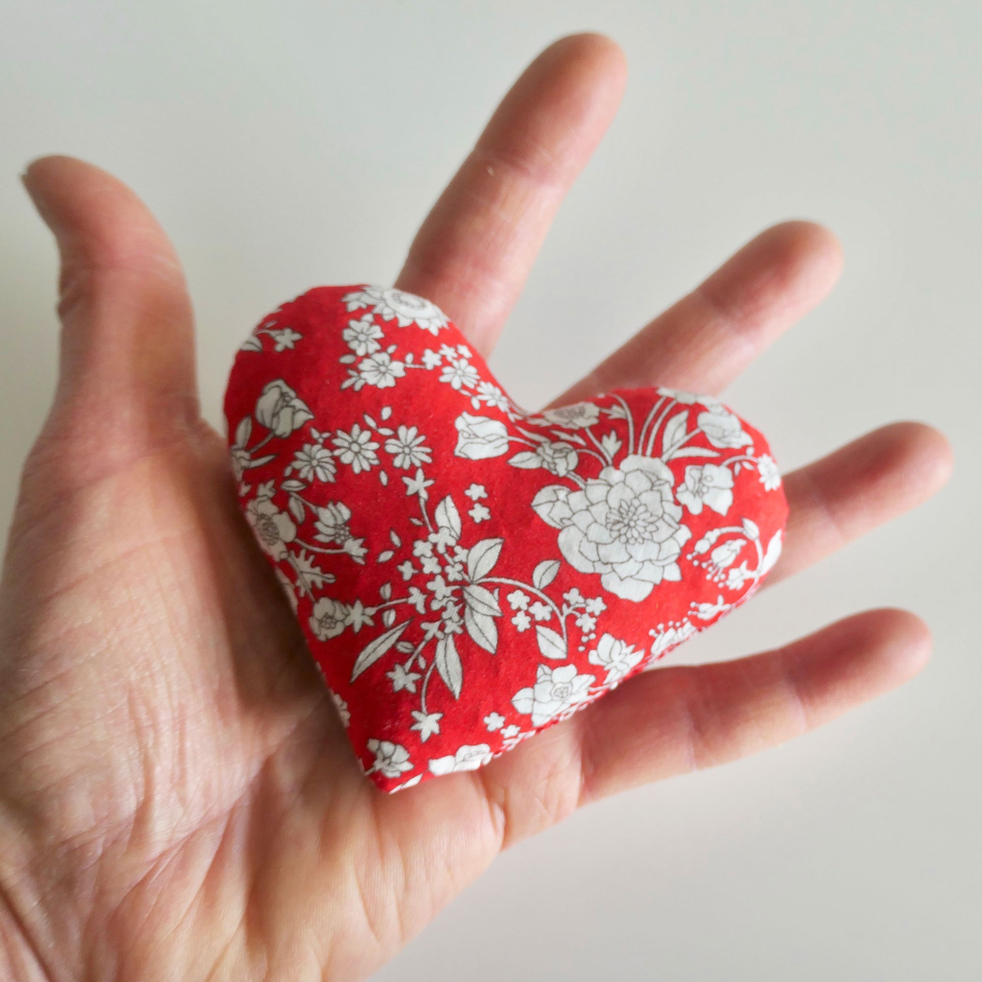 Hand holding a red heart-shaped fabric sachet with white floral pattern on a light background