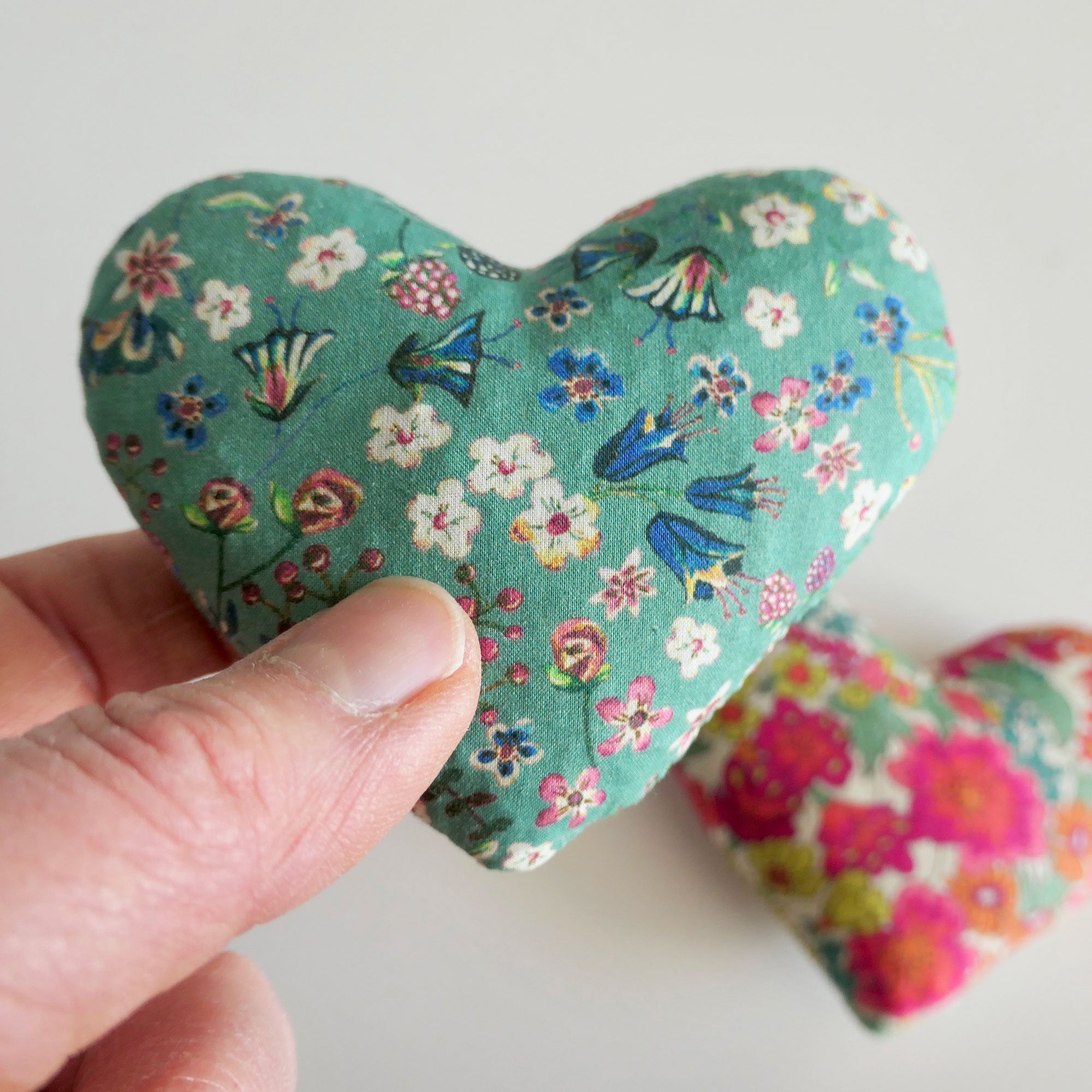 Floral-patterned heart-shaped fabric sachet held by a hand on a light background