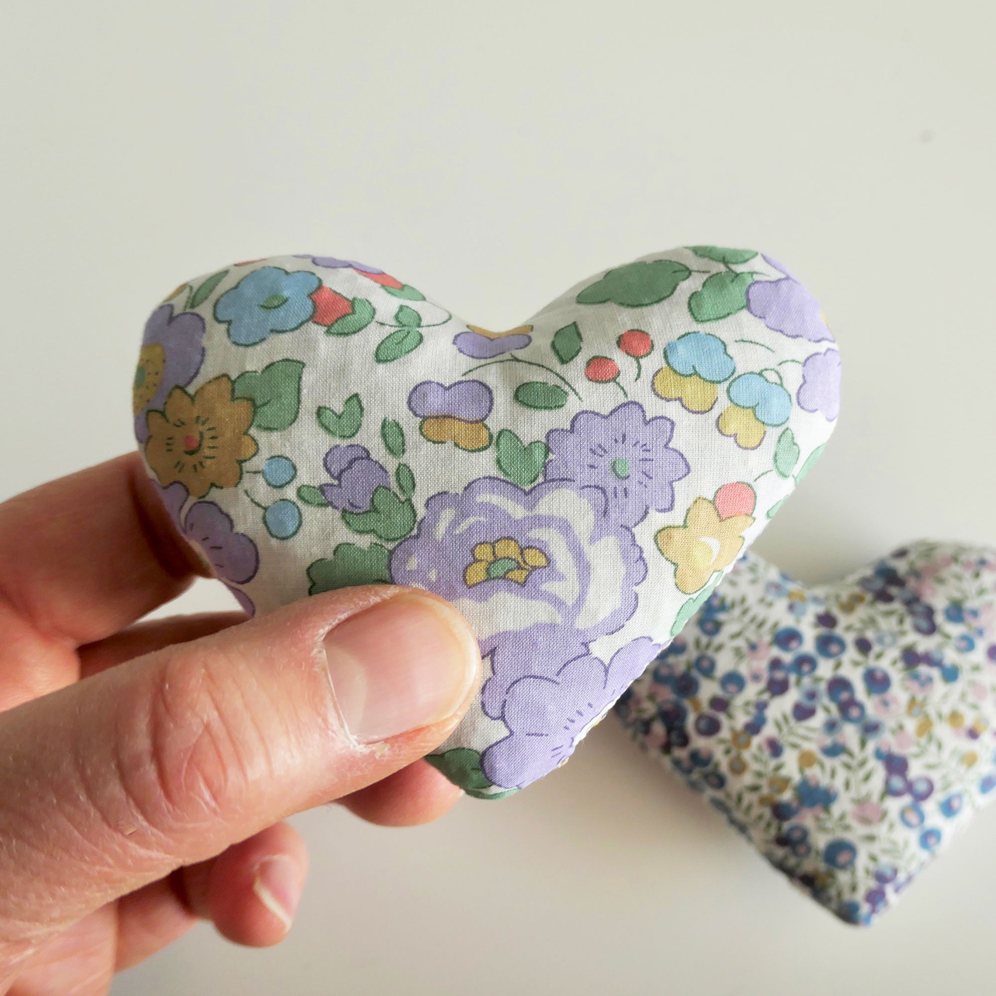 Hand holding a floral-patterned heart-shaped fabric sachet against a plain background