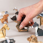 An image of a hand placing a dog into an arrangement of origami dogs on a neutral background.