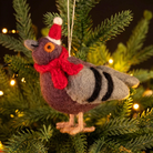 A felted wool Christmas ornament shaped like a pigeon, wearing a red and white Santa hat and a red scarf, hanging on a green Christmas tree with warm string lights.