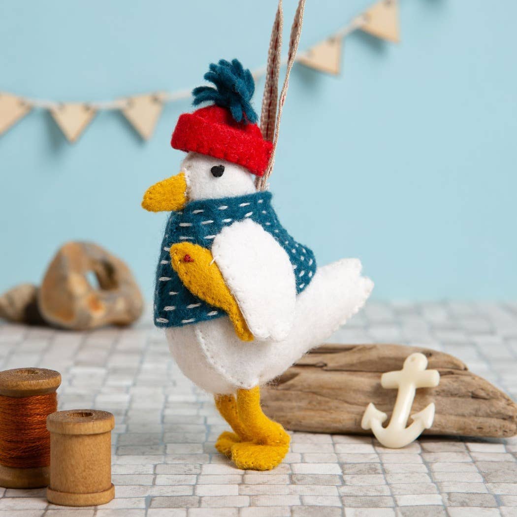 A white felt seagull toy wearing a red and blue hat, holding a yellow felt fish under their wing, surrounded by nautical items and colours.
