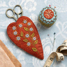 Embroidered red heart-shaped scissors pouch, colourful mini pincushion with botanical design, and sewing tools on a patterned blue fabric surface.