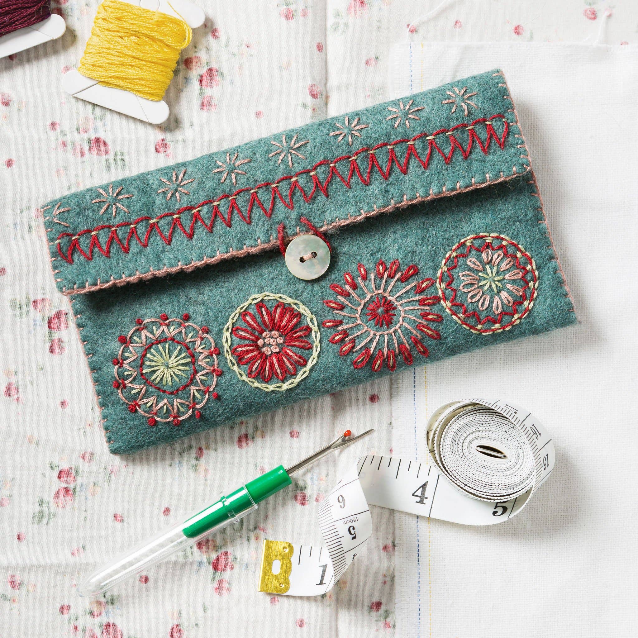 Image of light teal embroidered felt sewing pouch surrounded by sewing paraphernalia such as stitch unpicker, measuring tape, thread spools and fabric.