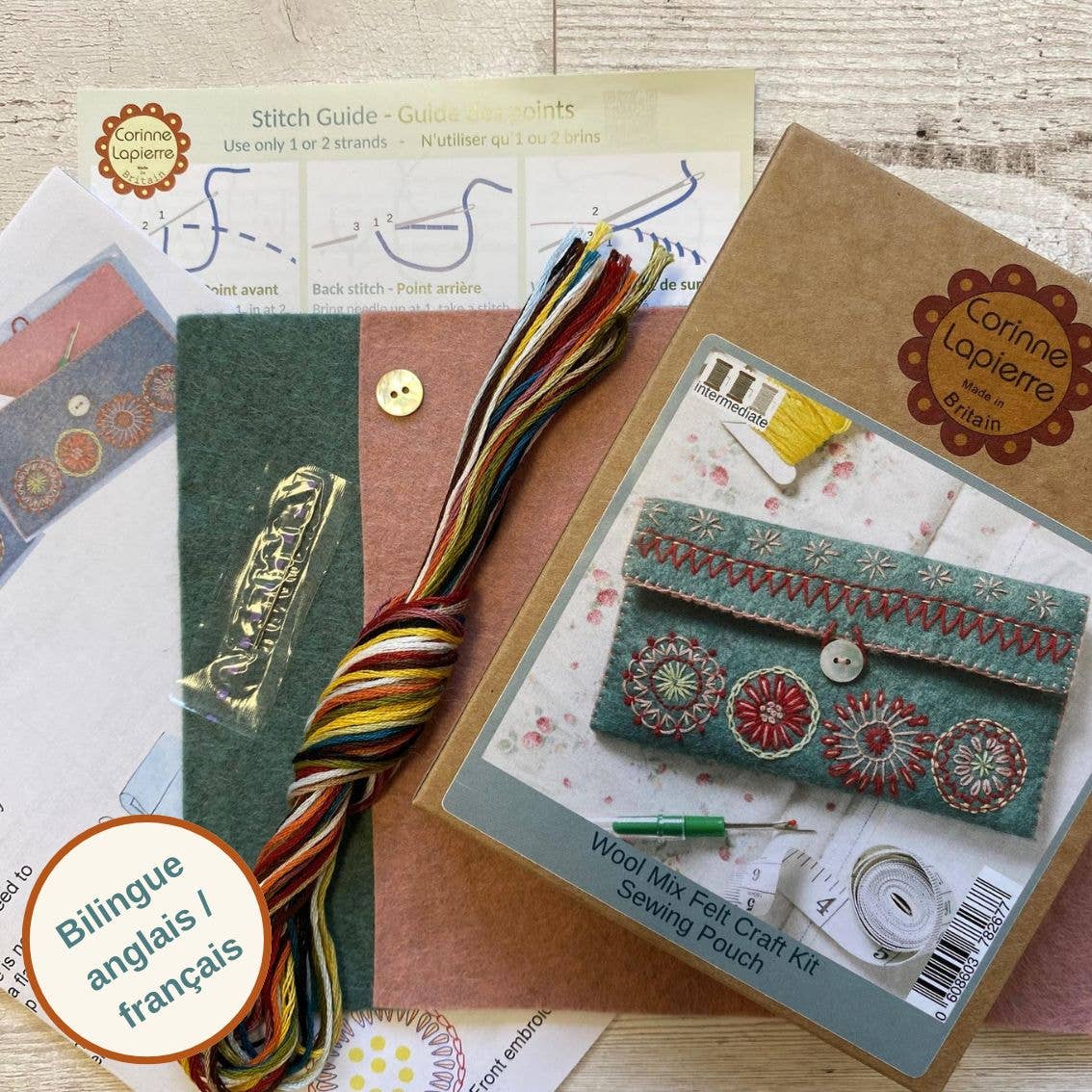 Brown kraft packaging box with photo of finished product on, an instruction sheet, colourful thread, pink and teal felt fabrics, a mother of pearl small round button and a needle laid out on a rustic wooden background.