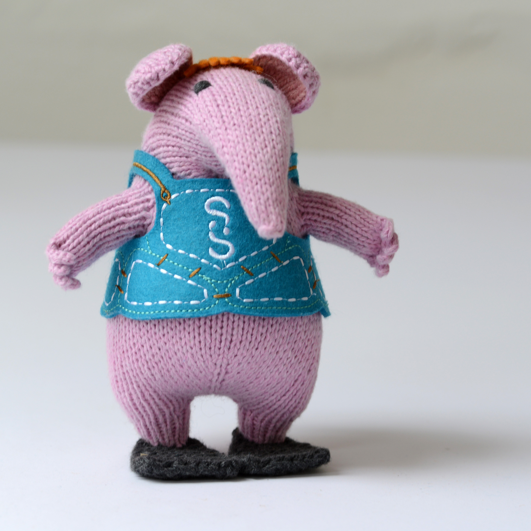 A knitted plush mouse character standing on a white surface against a light grey background. The plush has pink knit ears and legs, a blue vest with white embroidered stitching', and black shoes.
