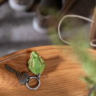 A green frog-shaped keychain with a metal keyring and attached keys, resting on a wooden surface with a softly blurred background.