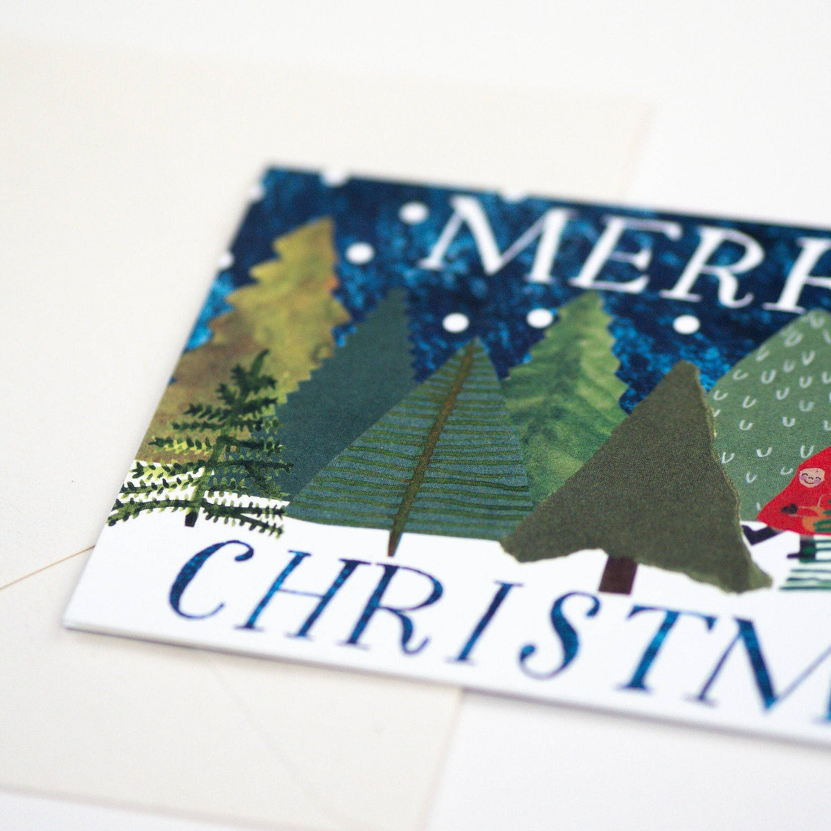 A close-up of a Christmas greeting card featuring a textured, collage-style winter forest with green and yellow trees under a dark blue sky, with the words 'MERRY CHRISTMAS' partially visible.