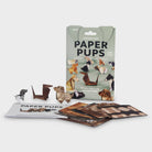 A boxed craft kit labeled 'PAPER PUPS' with several small origami dog figures and flat origami paper pieces arranged in front of the package on a light grey background.