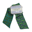 A green patterned necktie with blue and gray floral motifs, tied with a silver metal ribbon that has a white paper label reading 'LES BELLES VAGABONDES France Allemande'.