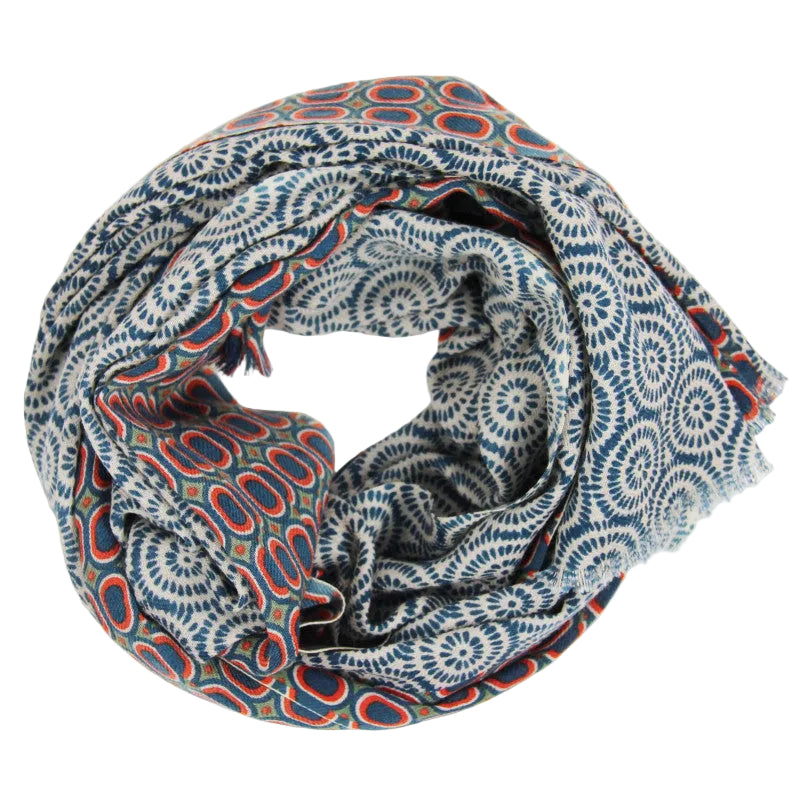 A lightweight scarf with a complex ikat-inspired pattern featuring blue, red, and white circular and paisley motifs, draped in a loose coil on a white background. 