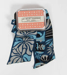 A patterned silk scarf in shades of blue, gray, and black, draped next to its silver metal tin packaging with a red and white label r